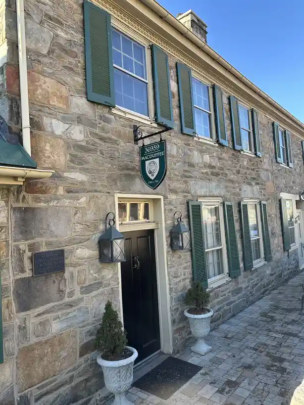 A stone building with green shutters and a black door has a green shield-shaped sign above the door that reads '36959…