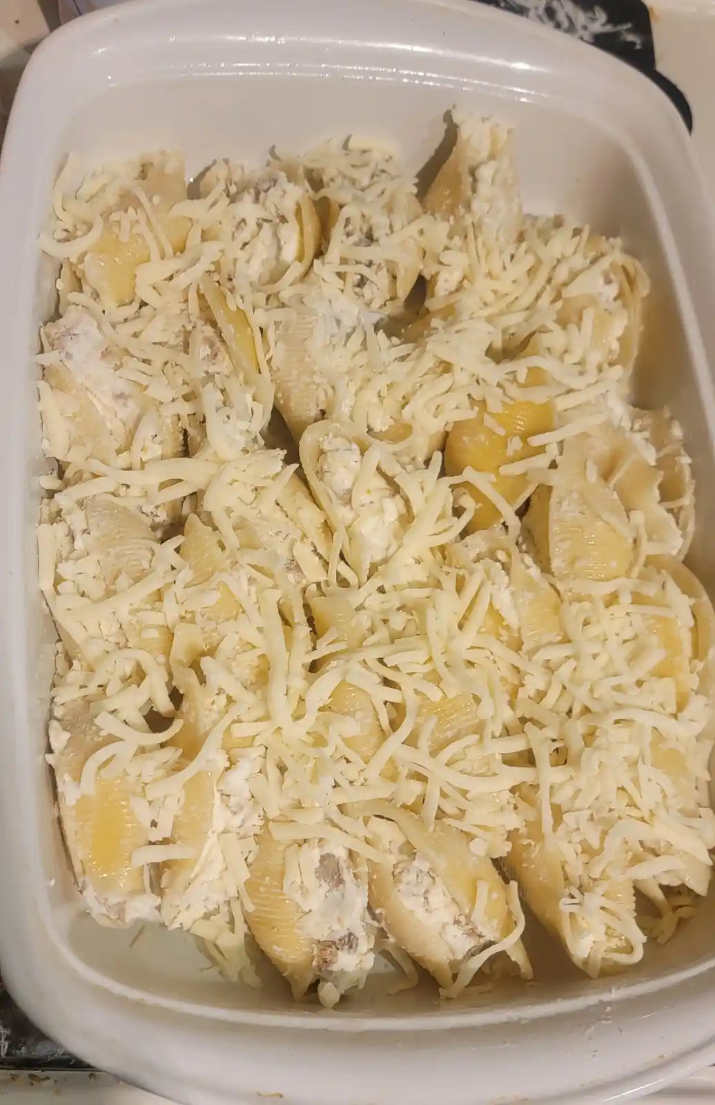 A white baking dish filled with uncooked pasta shells stuffed with a filling and generously topped with shredded cheese. The…
