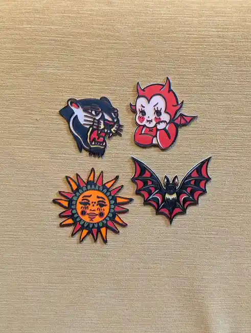 Four enamel pins are arranged on a textured beige surface: a snarling black panther, a cute red devil with horns and wings…