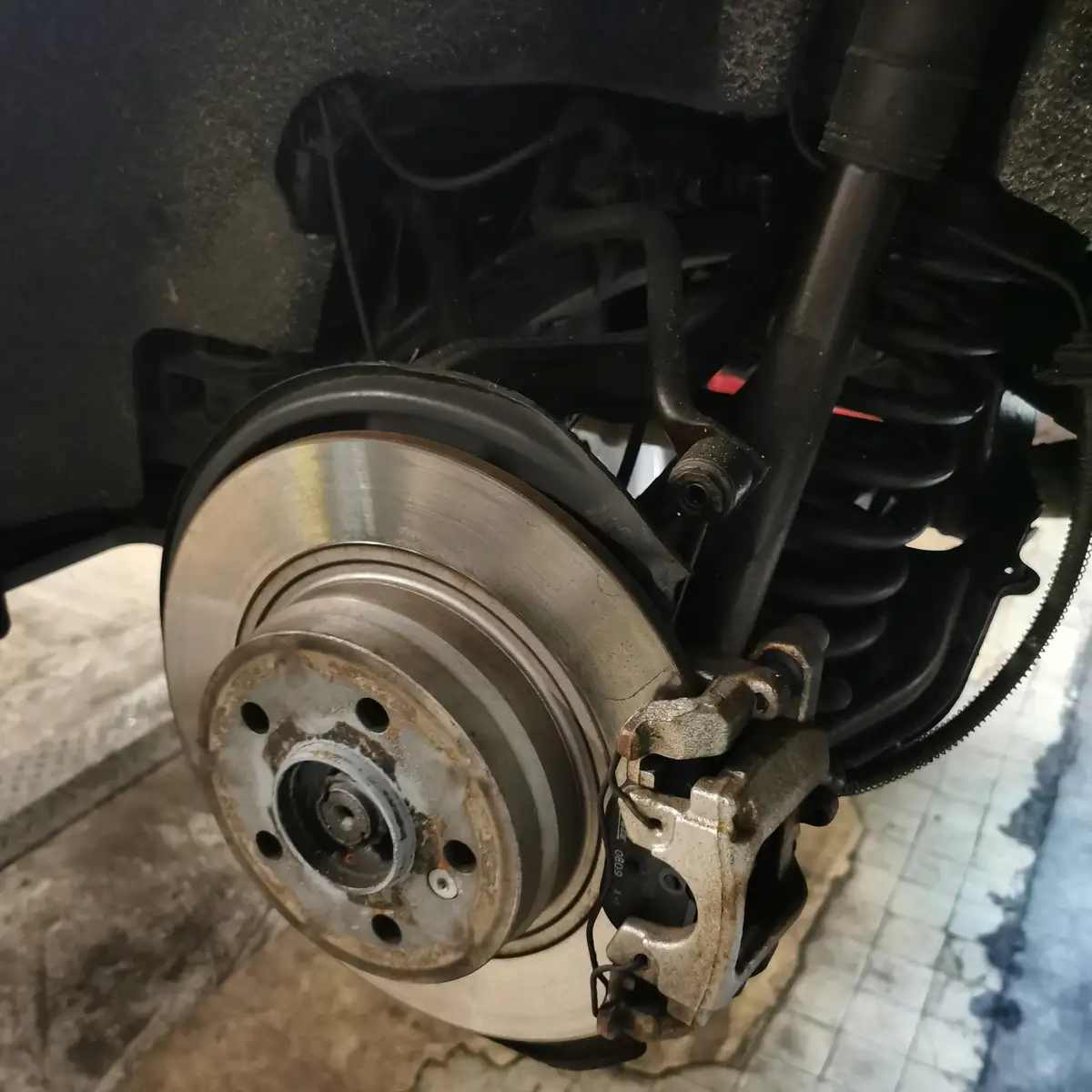 A close-up photo of a car's exposed brake system, showing a shiny metal brake rotor, a rusty brake caliper, and a coil…