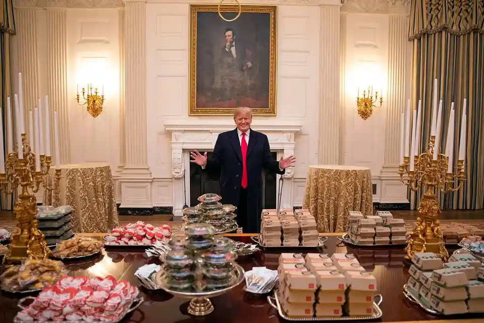 a man in a dark suit and red tie stands with arms outstretched behind a large table laden with various snack items including…