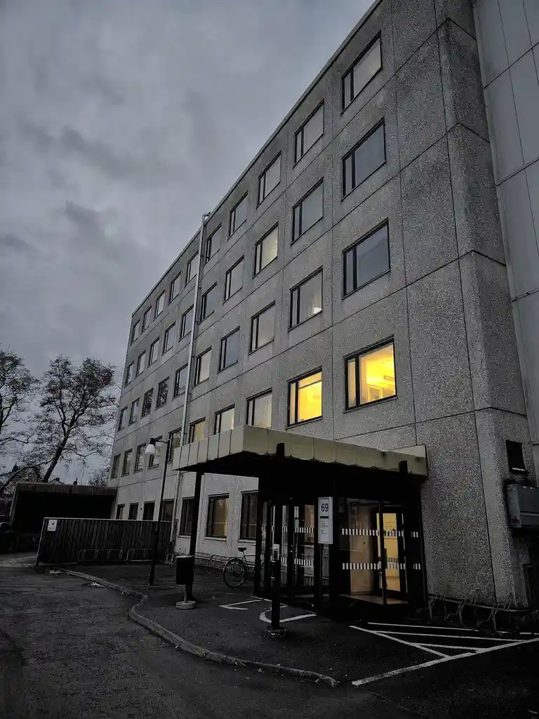 A low-angle shot of a grey, multi-story building with a cloudy sky, several windows are lit from within, and a bicycle is…