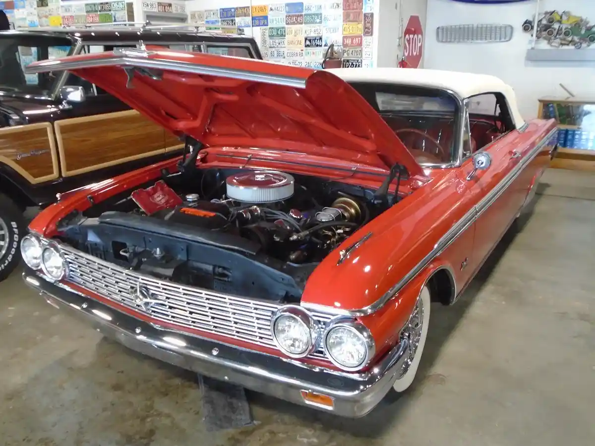 A red and white classic American car with its hood open, revealing a detailed engine, is parked indoors in a garage or…