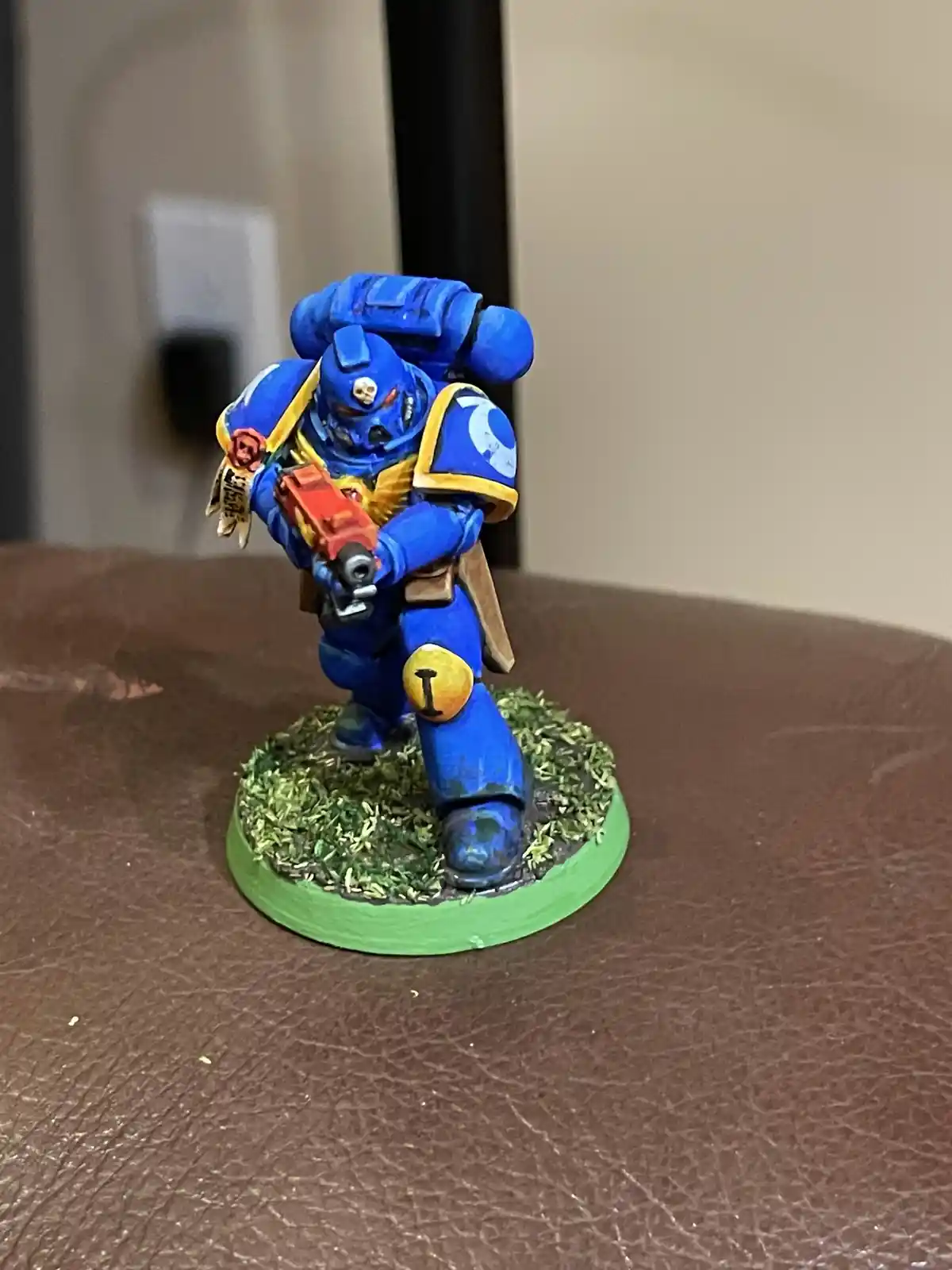 A blue-painted miniature of a Space Marine from Warhammer 40,000 stands on a green base with grass texture, holding a red…