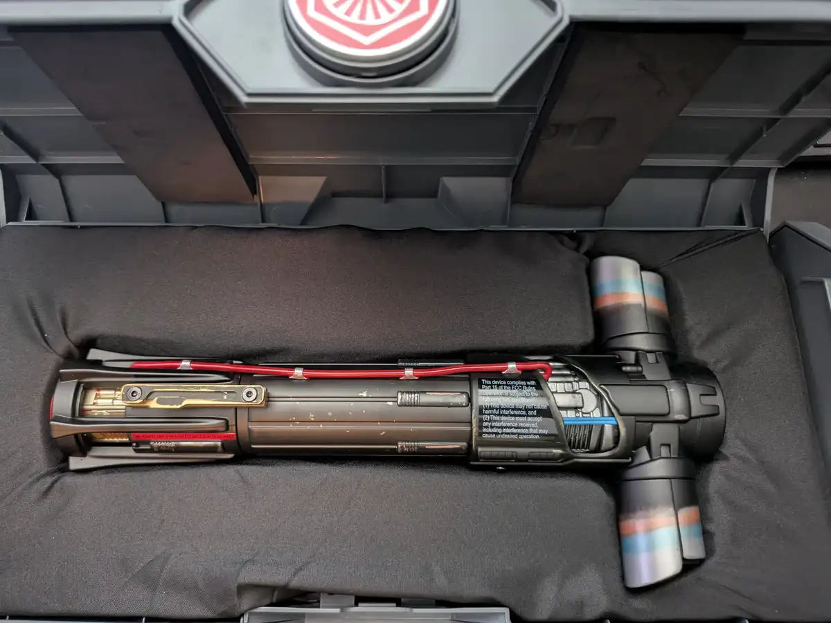 A custom-built lightsaber replica, resembling a Star Wars weapon, is shown lying horizontally inside a grey plastic storage…