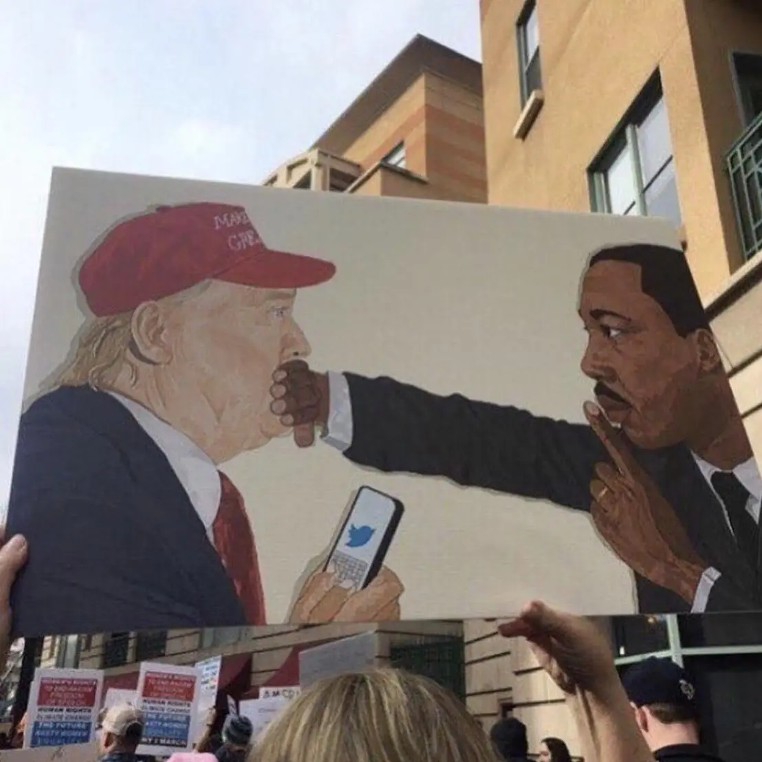 A large protest sign depicts a painted scene of Martin Luther King Jr. silencing Donald Trump by placing his finger over…