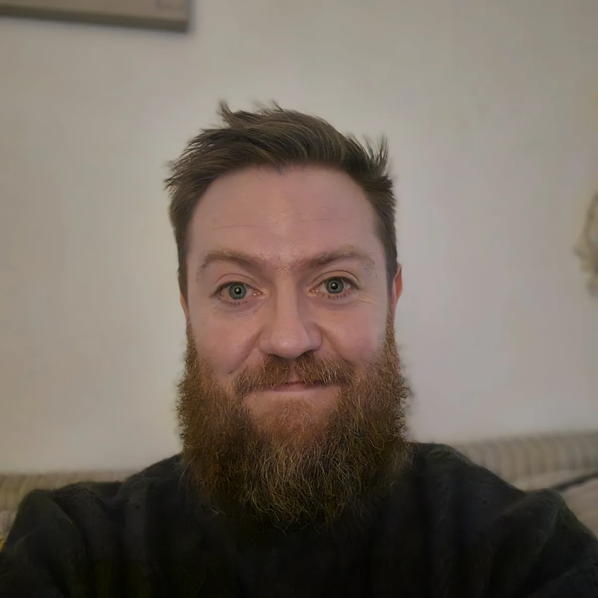 A man with a full reddish-brown beard and short brown hair looks directly at the camera with a tired, slightly annoyed…