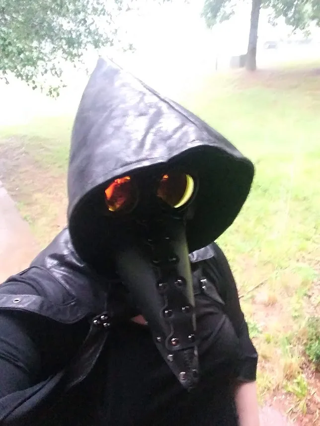A person wearing a black plague doctor mask with glowing orange eyes and a long beak, taking a selfie outdoors in a grassy…