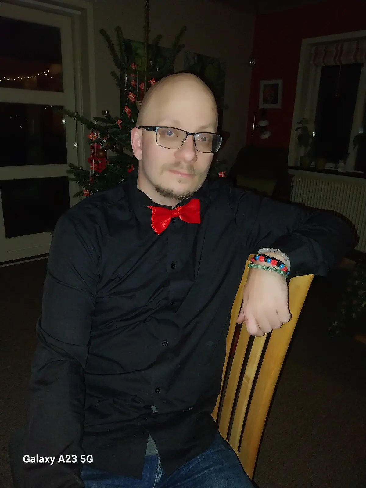 A bald man with glasses and a goatee sits in a wooden chair wearing a black button-up shirt and a bright red bow tie. He has…