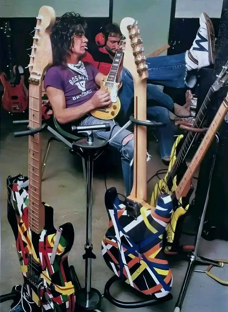 A man sits in a chair surrounded by multiple electric guitars, some with colorful abstract designs, while casually resting…