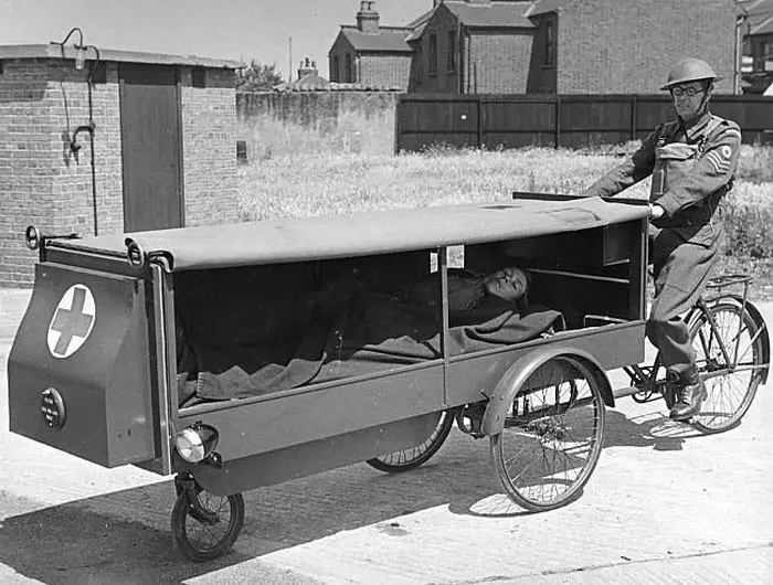 A black-and-white photo shows a soldier in uniform pedaling a three-wheeled military ambulance cart with a red cross emblem…