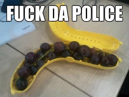 A yellow banana-shaped fruit holder filled with dark purple grapes sits on a wooden desk, with the text 'FUCK DA POLICE'…