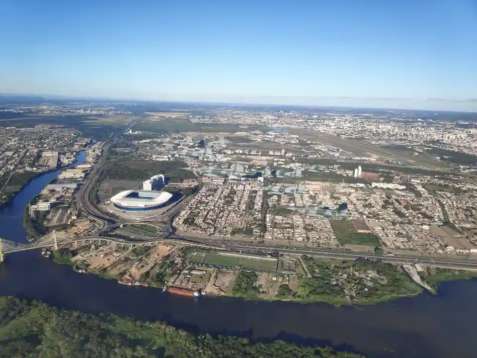 An aerial photograph shows a sprawling cityscape with a large river curving through it, a prominent modern stadium with a…