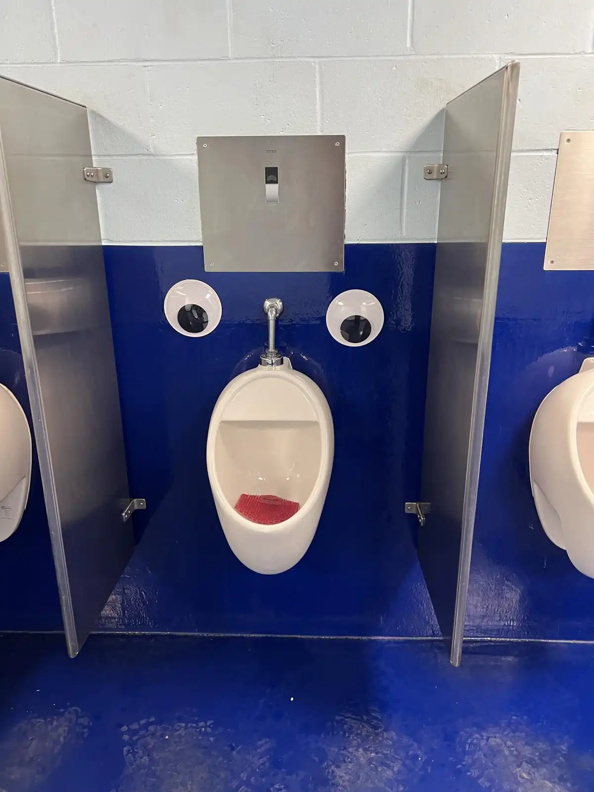 A white urinal in a public restroom stall with a blue wall has two large googly eyes glued to the wall on either side of it…