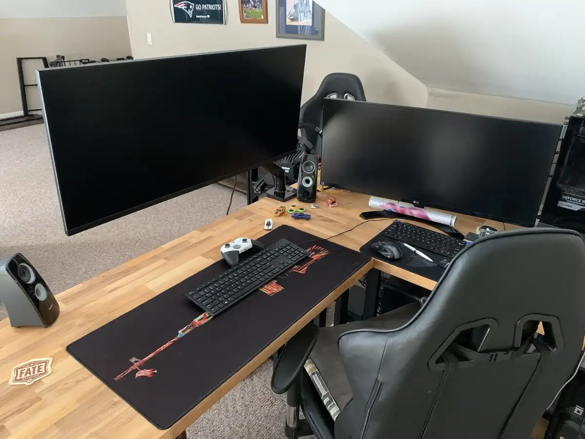 A photo of a gaming setup with two large, black, turned-off monitors on a wooden desk, a black gaming chair, a keyboard, a…