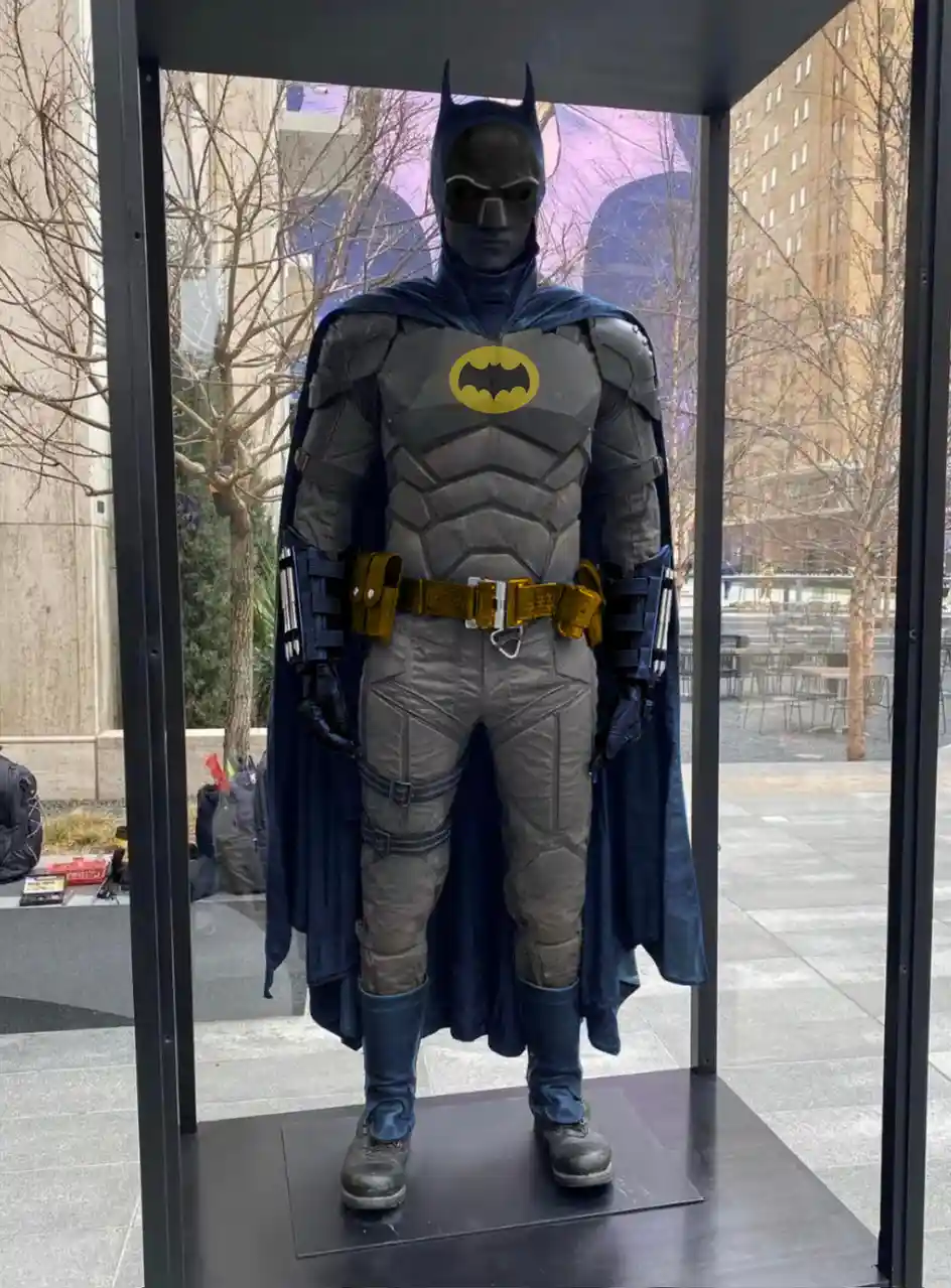 A full-body Batman costume, resembling the armored suit from 'The Dark Knight Rises', is displayed in a glass case outdoors…