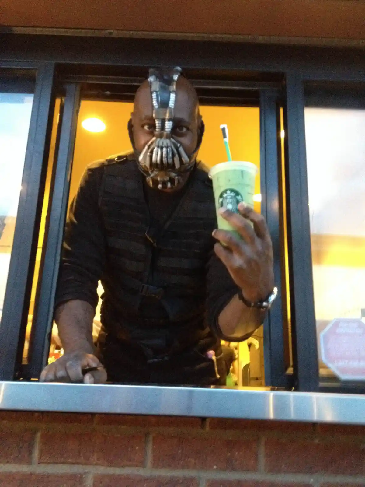 A person wearing a Bane mask and tactical vest leans out of a Starbucks drive-thru window, holding up a green Starbucks…