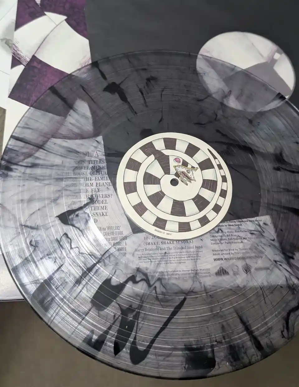 A close-up photo of a black and white marbled vinyl record, showing its label with a cartoon snake head and tracklist for…
