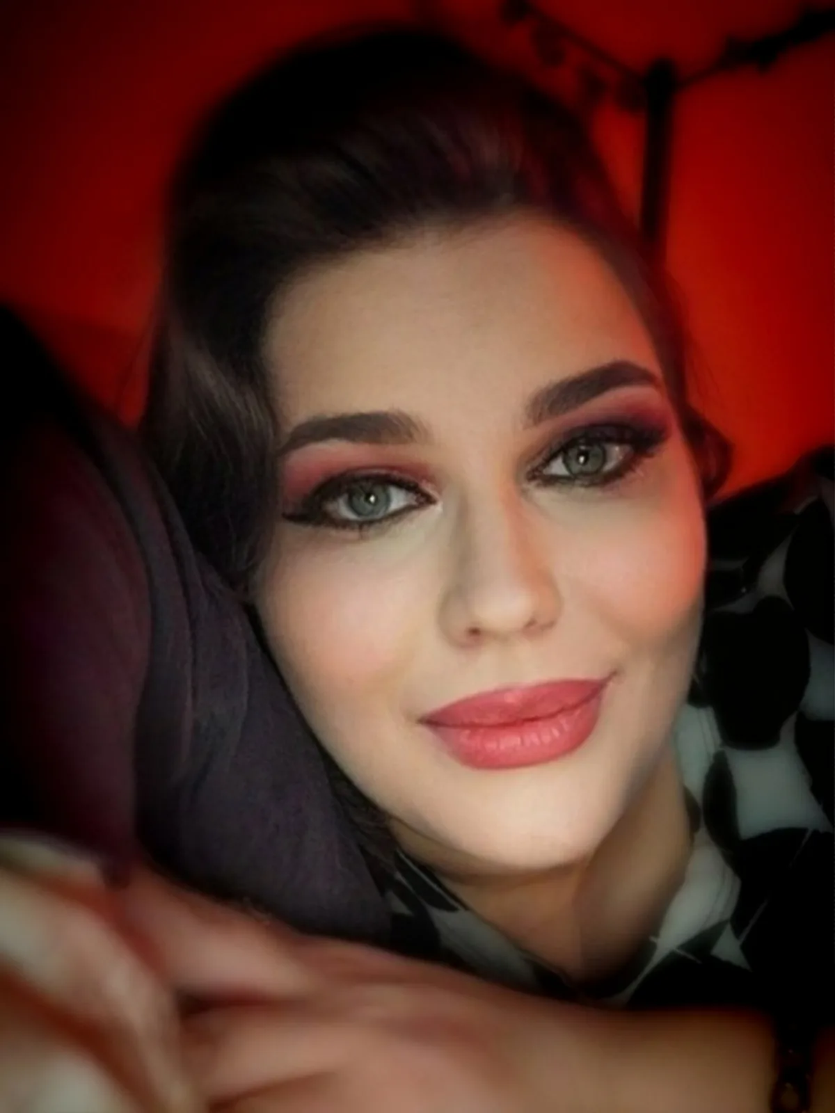A close-up selfie of a woman with dramatic makeup, including smoky eyeshadow and pink lipstick, smiling while lying down…