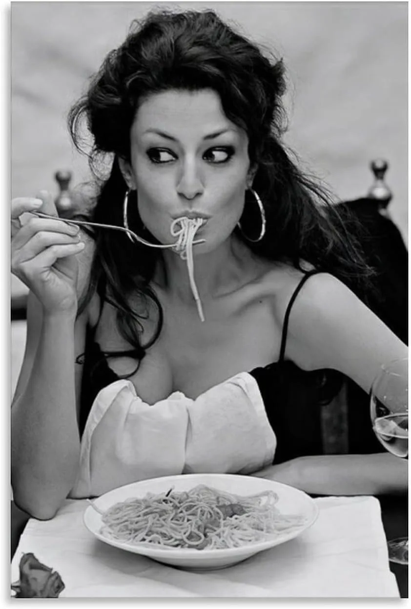 A black-and-white photo shows a woman with dark, wavy hair and large hoop earrings eating spaghetti from a fork, with…