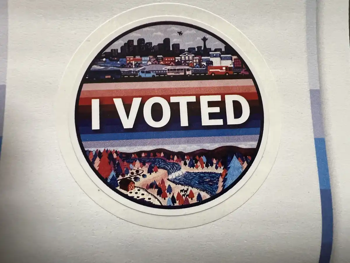 A circular 'I VOTED' sticker is affixed to a white surface, likely a shirt. The sticker features a stylized, colorful…
