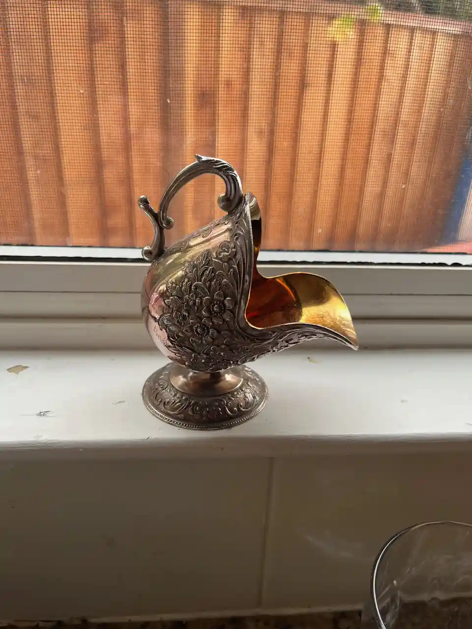 A silver-colored, ornate pitcher with a floral embossed pattern and a golden interior sits on a white windowsill, with a…