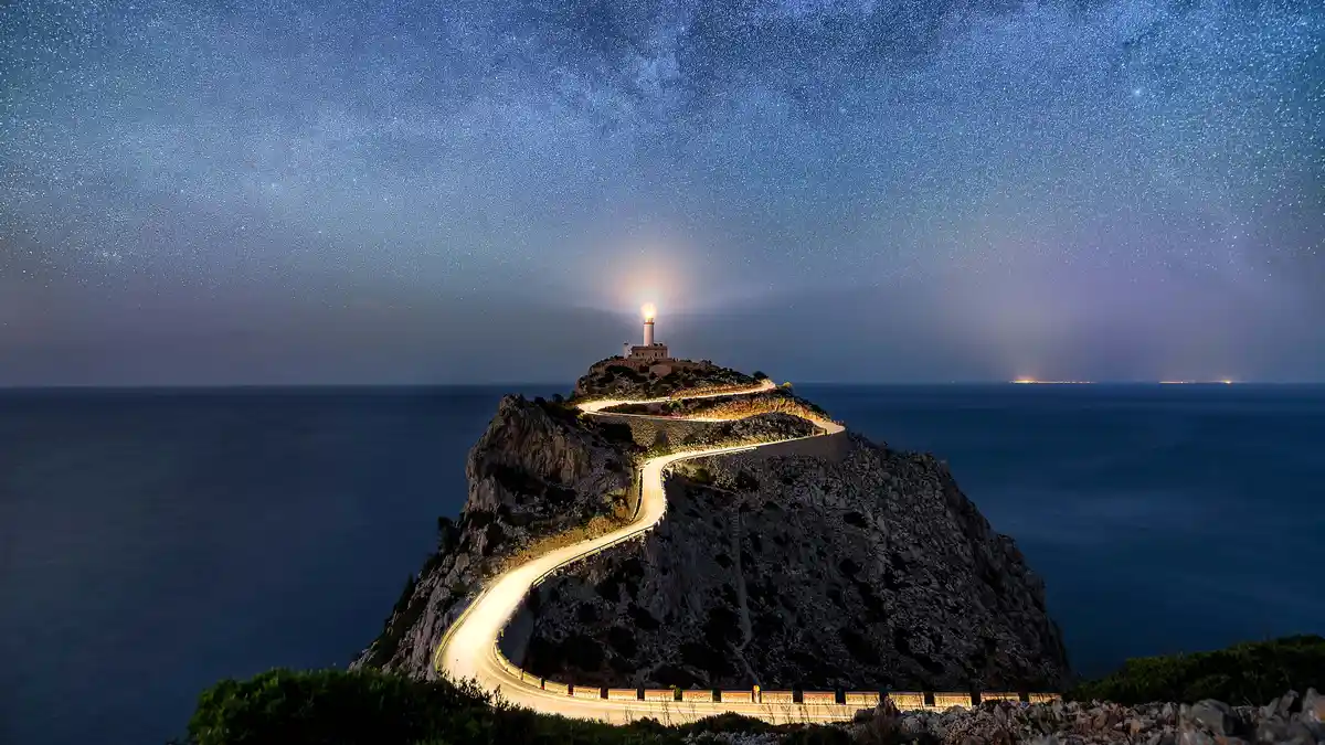 In this long exposure photo, snaking car light trails merge with the 1893 Formentor Lighthouse's shine, as stars illuminate…