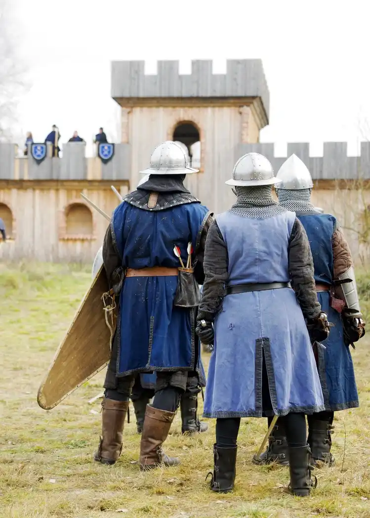 Three medieval reenactors in chainmail and helmets stand with their backs to the camera, facing a wooden castle with people…