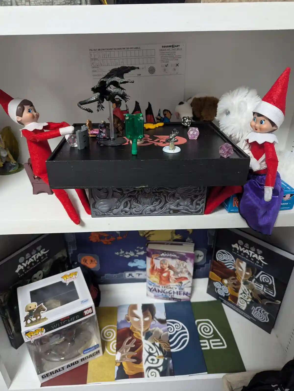 Two elf dolls in red and white outfits are positioned on a white shelf, appearing to play a tabletop role-playing game on a…