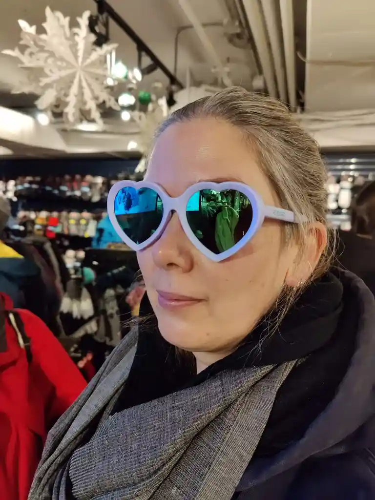 A woman with gray hair pulled back wears large, light purple-framed heart-shaped sunglasses with reflective lenses. She is…