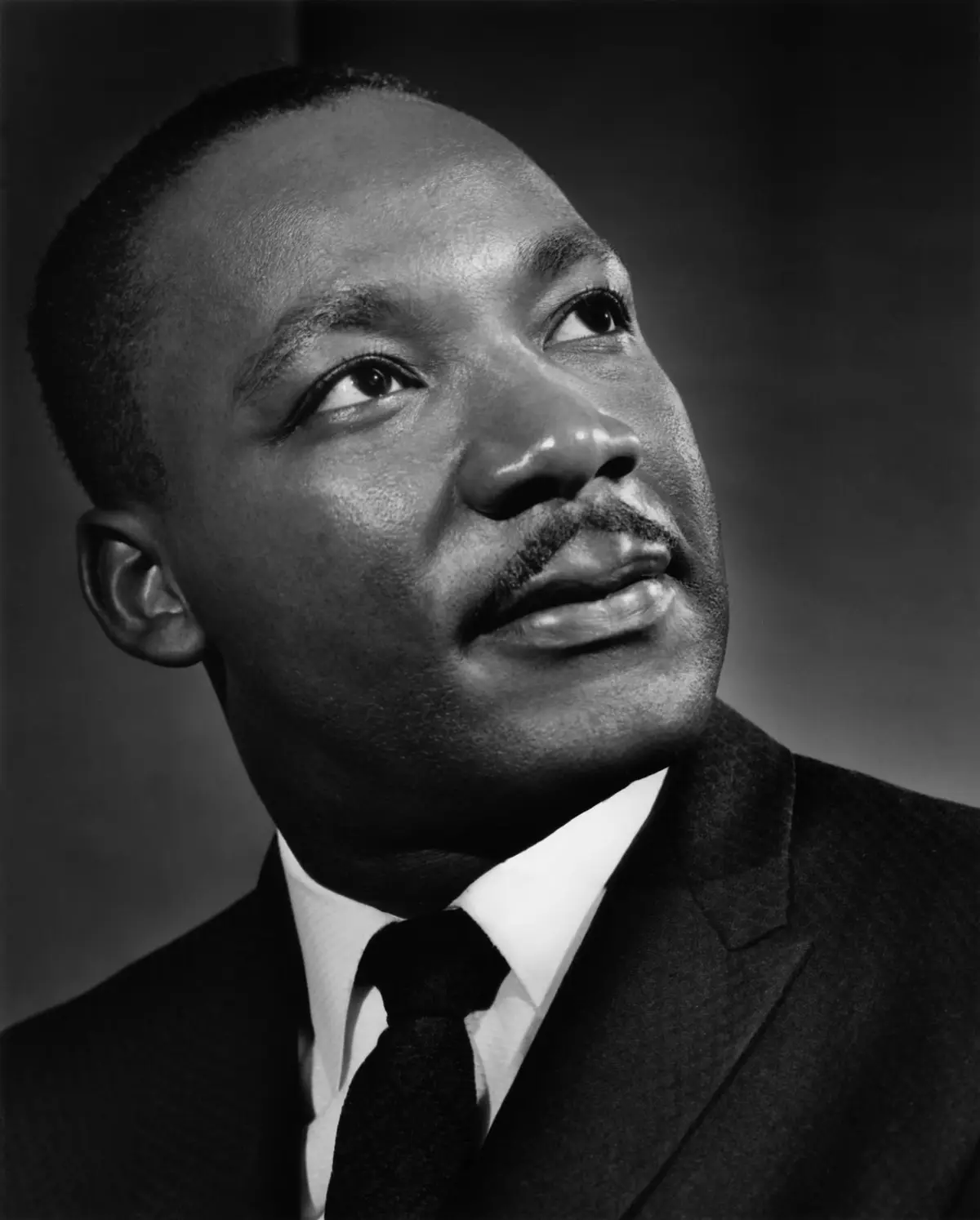 A black and white portrait of Martin Luther King Jr. looking upward with a thoughtful expression, overlaid with the text…