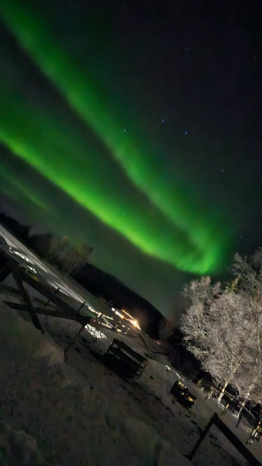 A nighttime photo shows vibrant green auroras dancing across a dark sky above a snowy landscape with trees and a wooden…