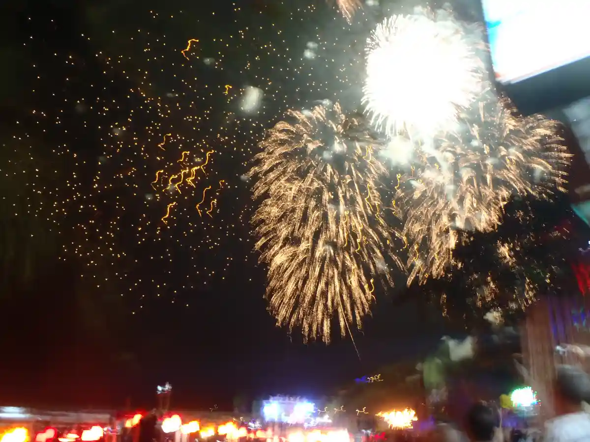 A blurry nighttime photo captures a large, bright fireworks explosion in the sky above a crowd, with golden sparks and…