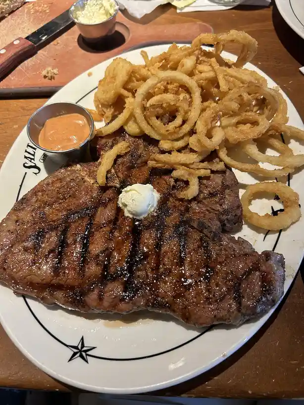 A close-up photo of a large, grilled steak on a white plate with black decorative lines and a star, topped with a pat of…