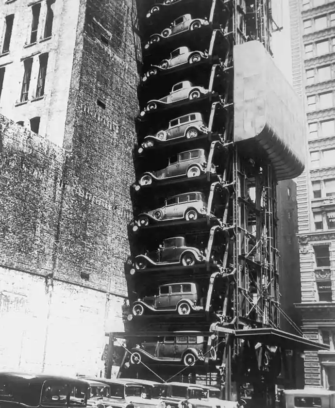 A black-and-white historical photo shows a tall, narrow, multi-level outdoor car parking structure attached to a brick…