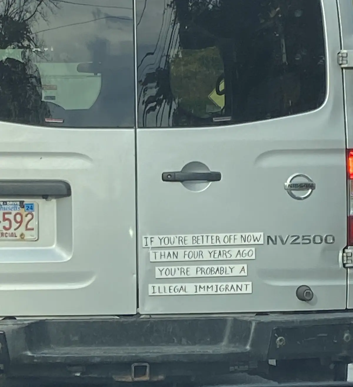 A close-up of the rear side of a silver Nissan NV2500 van shows four white rectangular stickers arranged vertically on the…