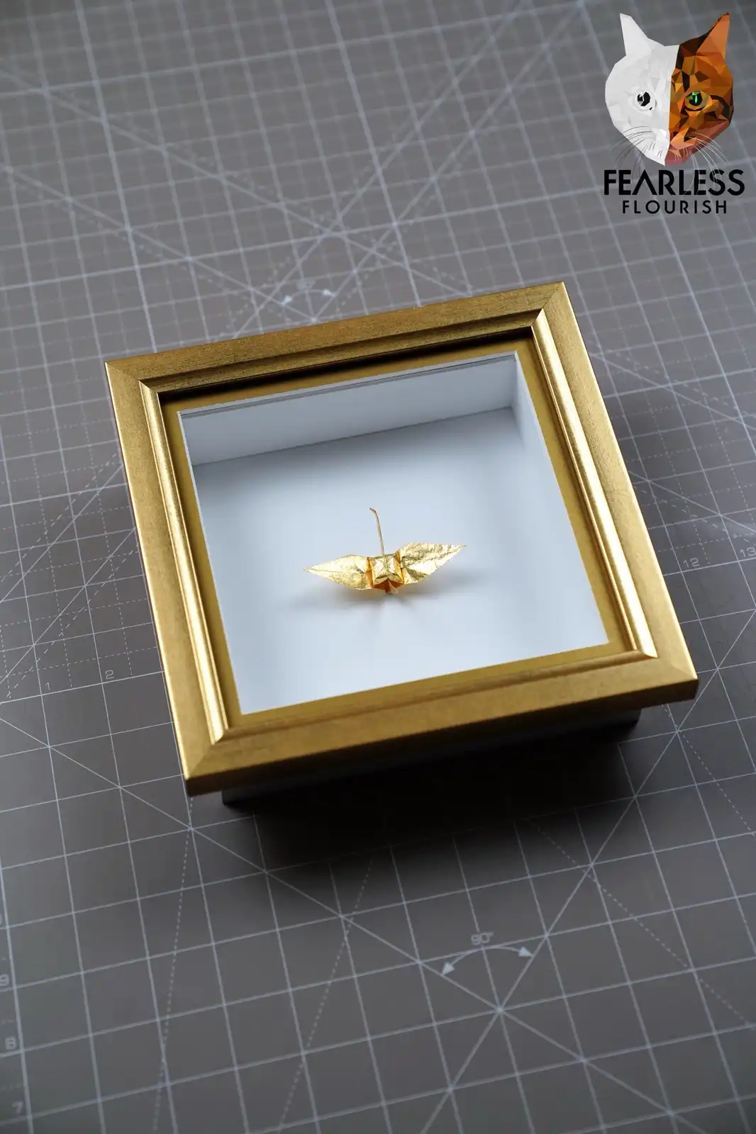 A small golden origami crane is displayed inside a square gold-colored shadow box frame, resting on a white backing. The…