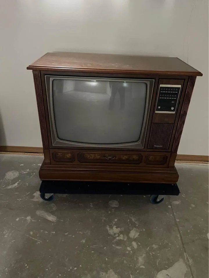A vintage wooden-cased Panasonic television with a blank screen sits on a black wheeled dolly on a concrete floor. The TV…
