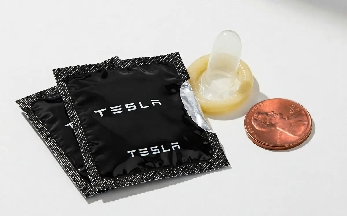A studio shot displays two black, foil-wrapped packages with the Tesla logo, next to a used condom and a US penny for scale…