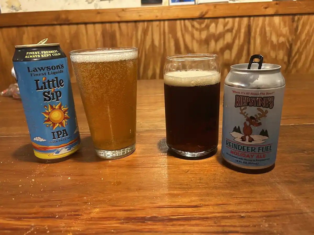Two beer cans and two glasses of beer sit on a wooden table. The can on the left is 'Lawson's Finest Liquids Little Sip IPA'…