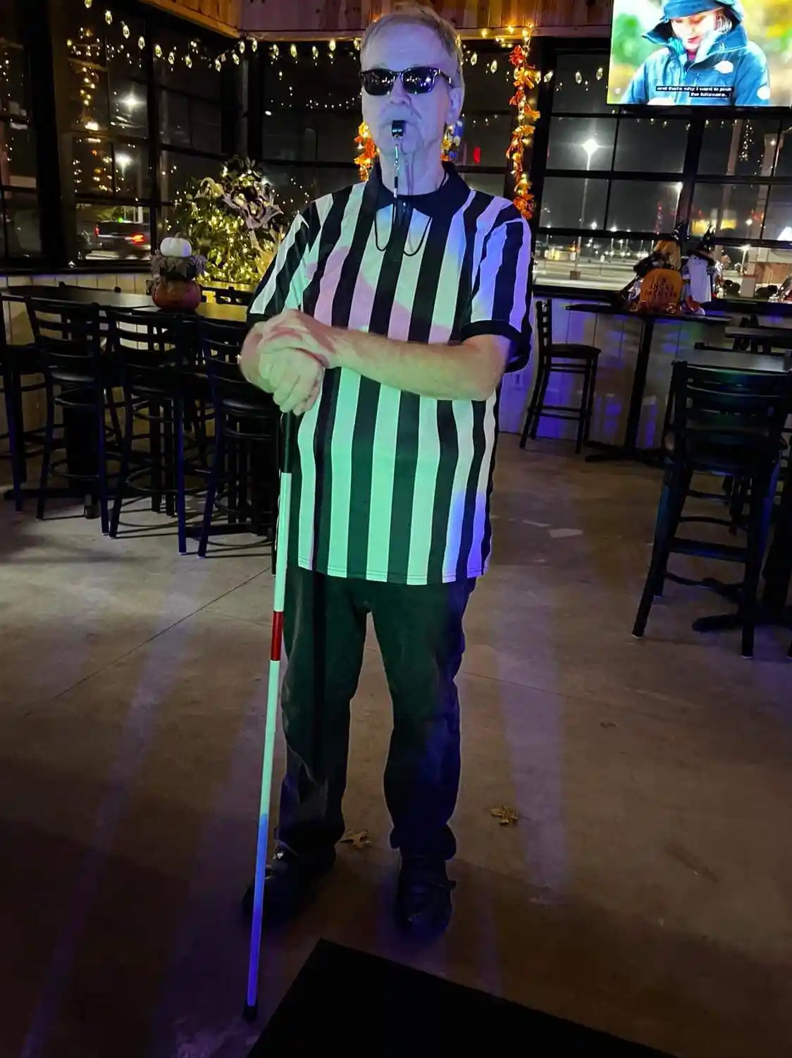 A man in a black-and-white striped referee shirt and sunglasses stands indoors holding a white cane with red and blue…