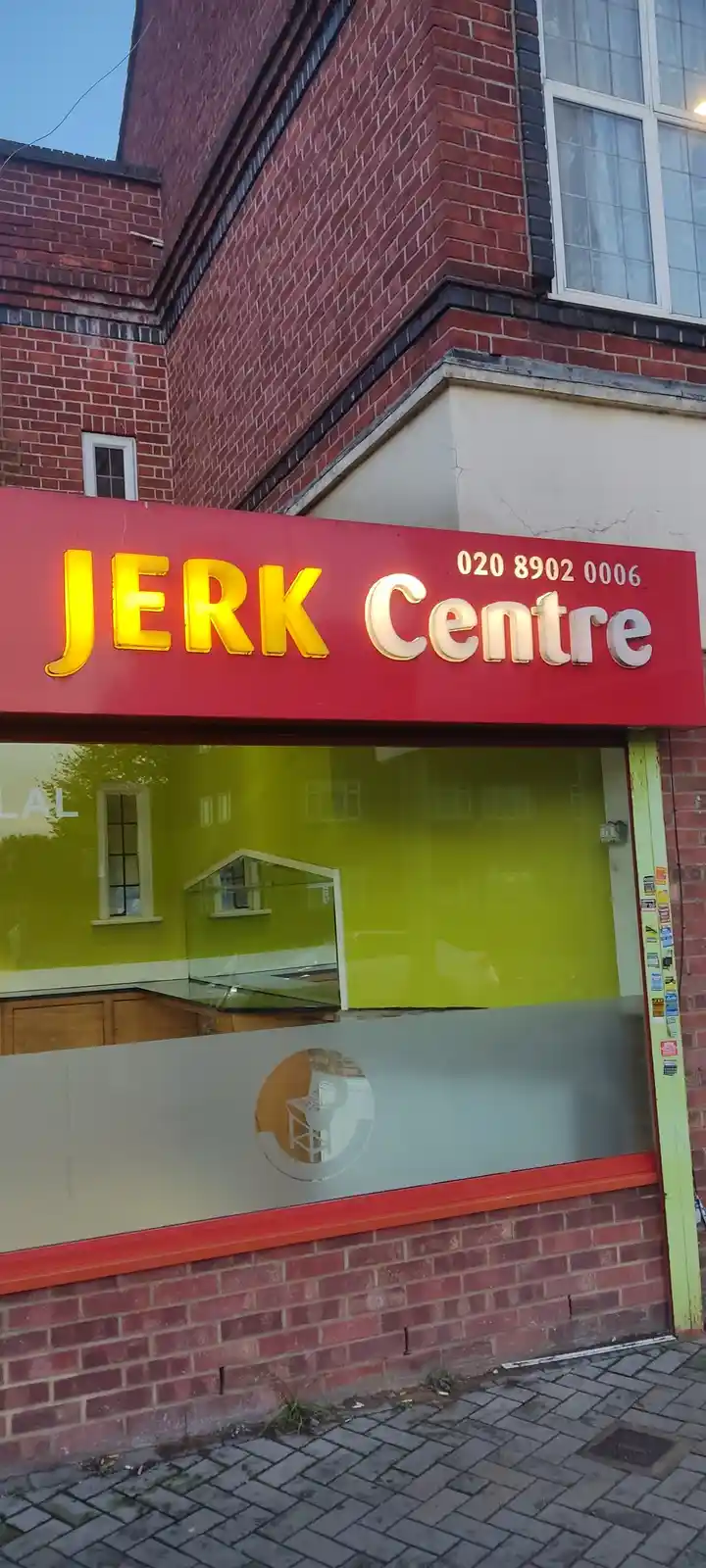 A storefront with a red sign that reads 'JERK Centre' in large yellow and white letters, with a phone number '020 8902 0006'…