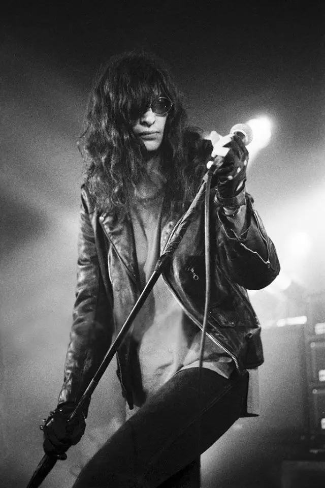 A black-and-white photo of a long-haired rock musician wearing sunglasses and a leather jacket, holding a microphone and a…