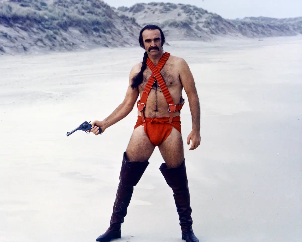 A man with a mustache and long dark hair in a braid stands in a desert-like landscape wearing a red loincloth, red harness…