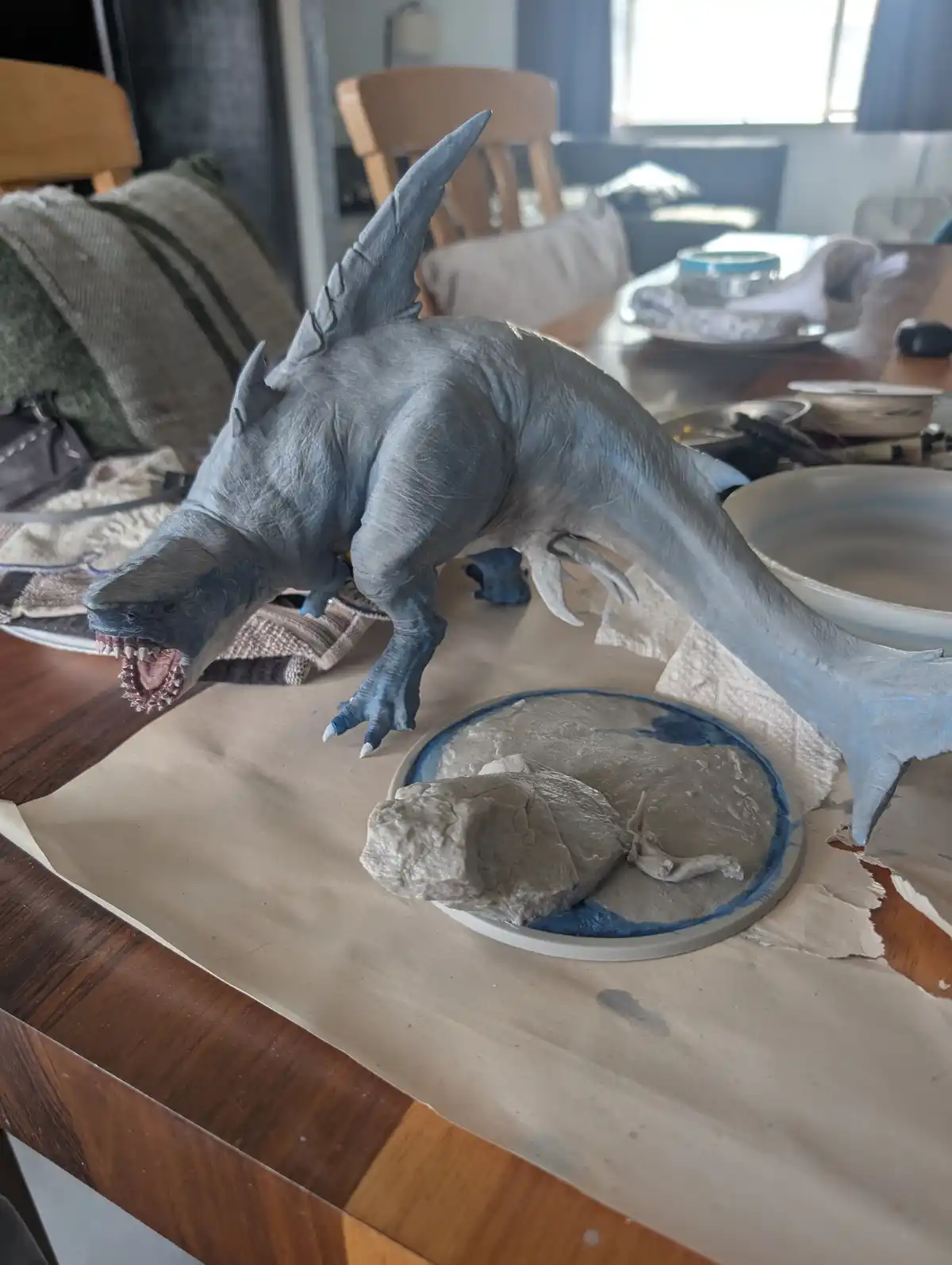 A grey, shark-headed, dinosaur-like creature sculpture with sharp teeth and claws is posed on a wooden table, leaning over a…