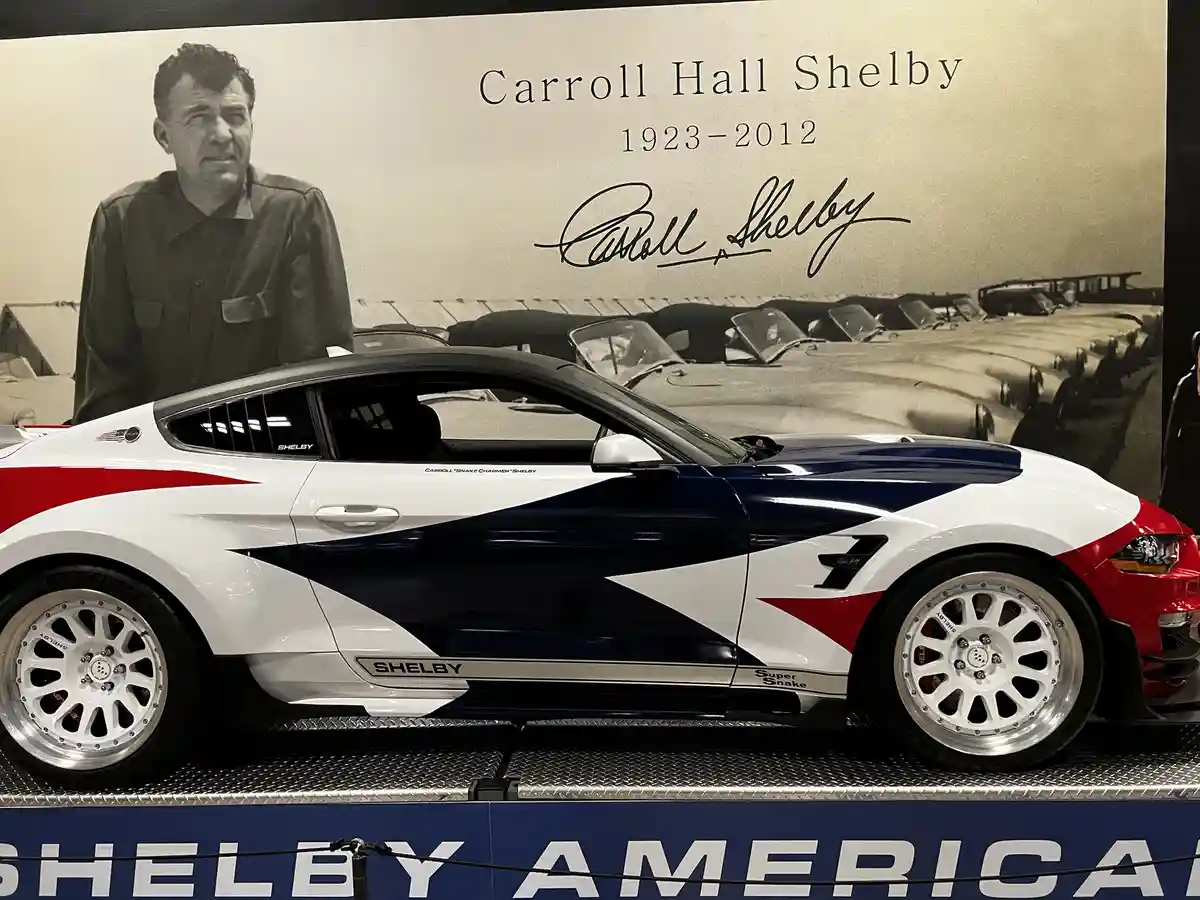A white, red, and blue Shelby Super Snake Mustang is displayed in front of a large black-and-white memorial wall featuring a…