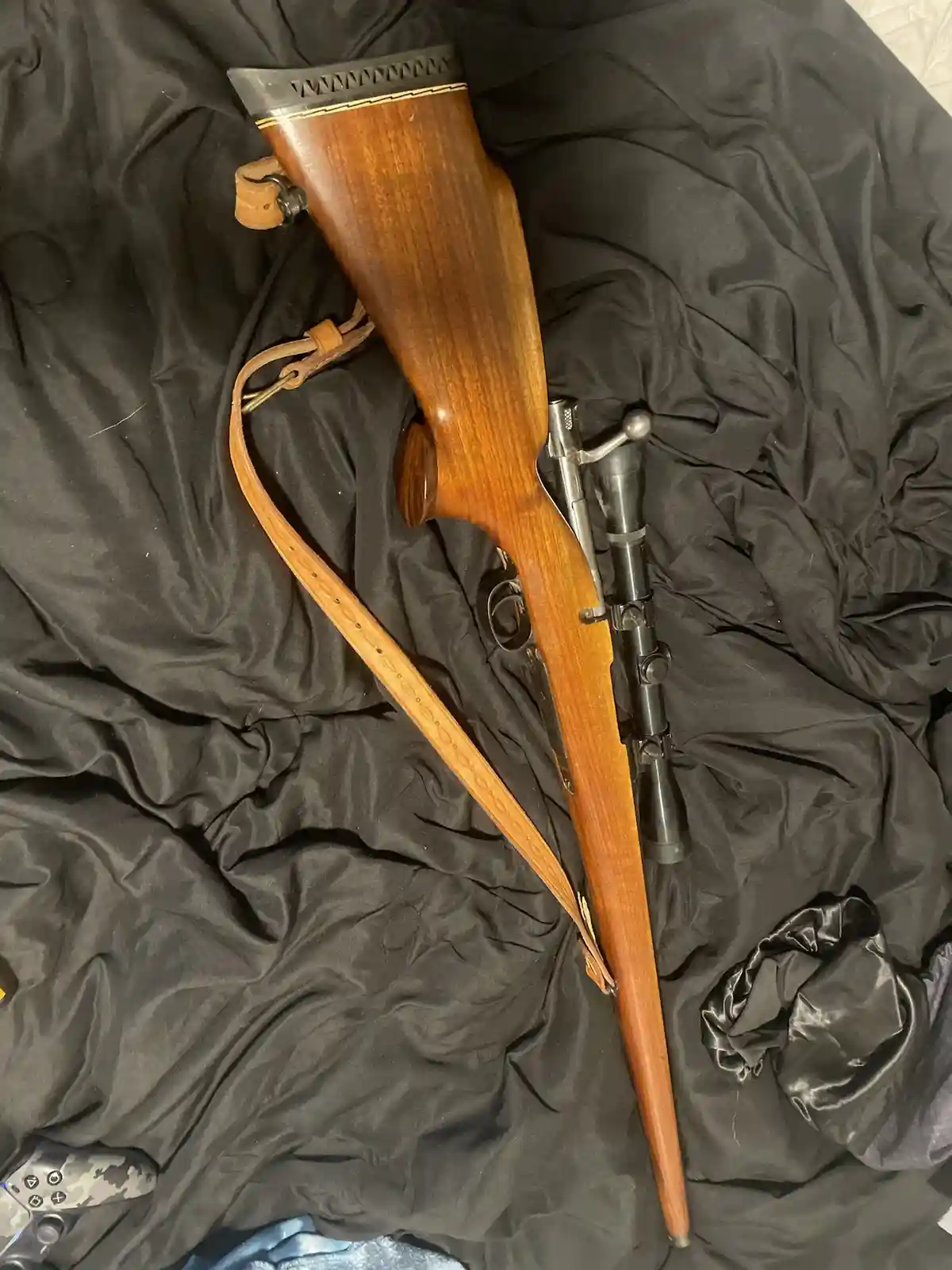 A wooden-stocked rifle with a scope and leather sling rests diagonally on a black fabric surface, next to a…
