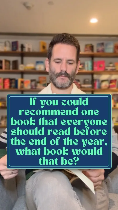 A man with a beard and mustache sits in a bookstore, looking down thoughtfully while holding a book. A large blue text box…