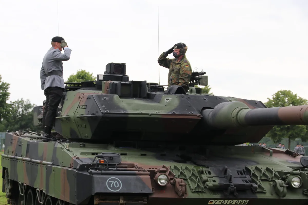 Two men in military uniforms stand atop a camouflaged tank, both saluting. One wears a grey uniform and beret, the other a…