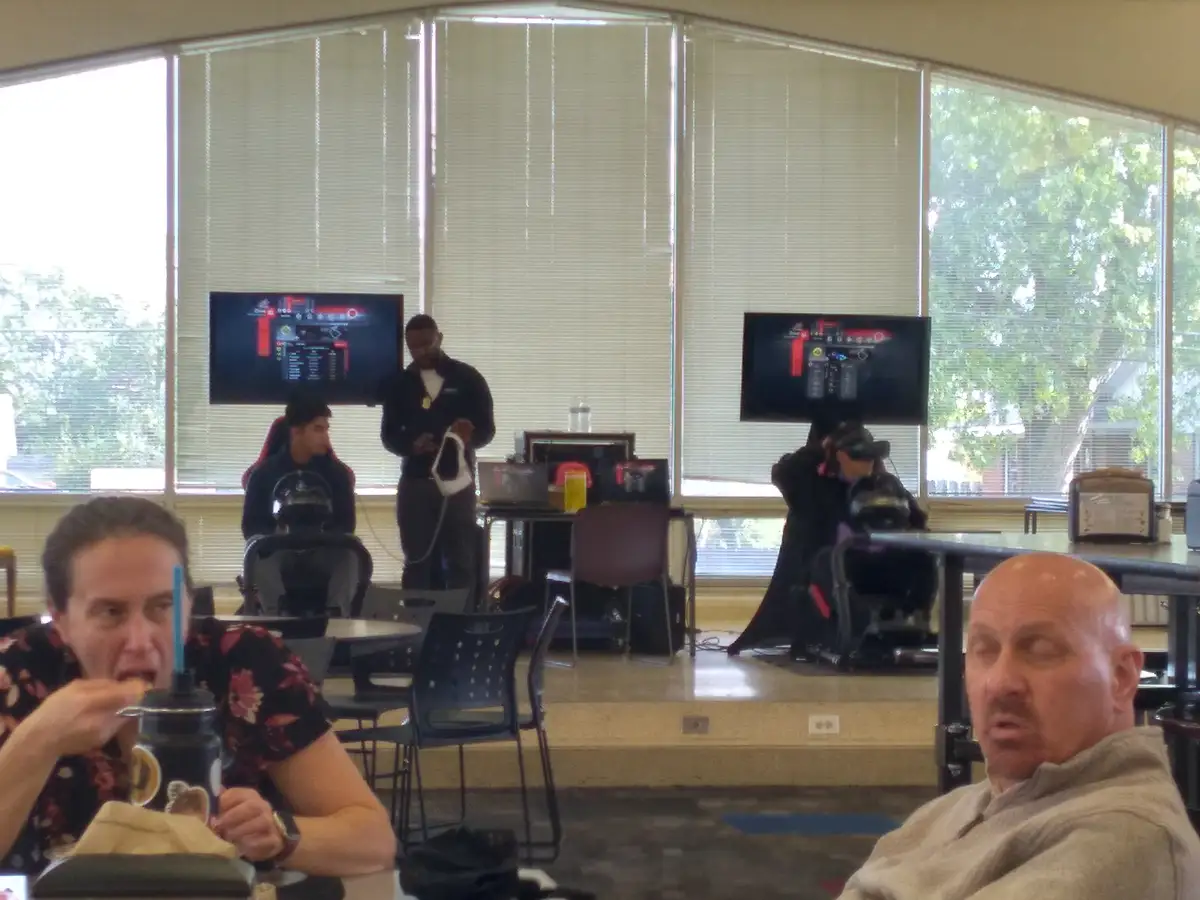 In a casual indoor setting with large windows and blinds, two people sit in the foreground: one on the left eating with a…